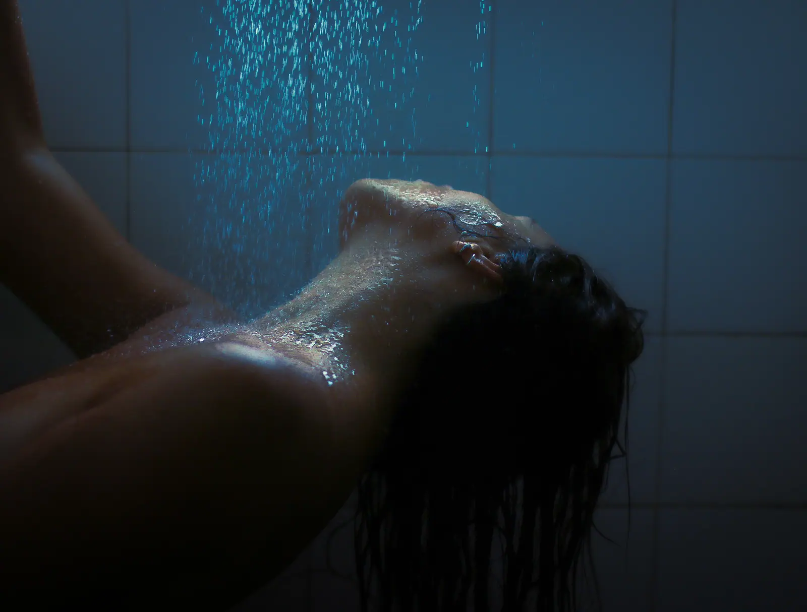 Woman under a shower, head tilted back, water lit blue against pale tiles